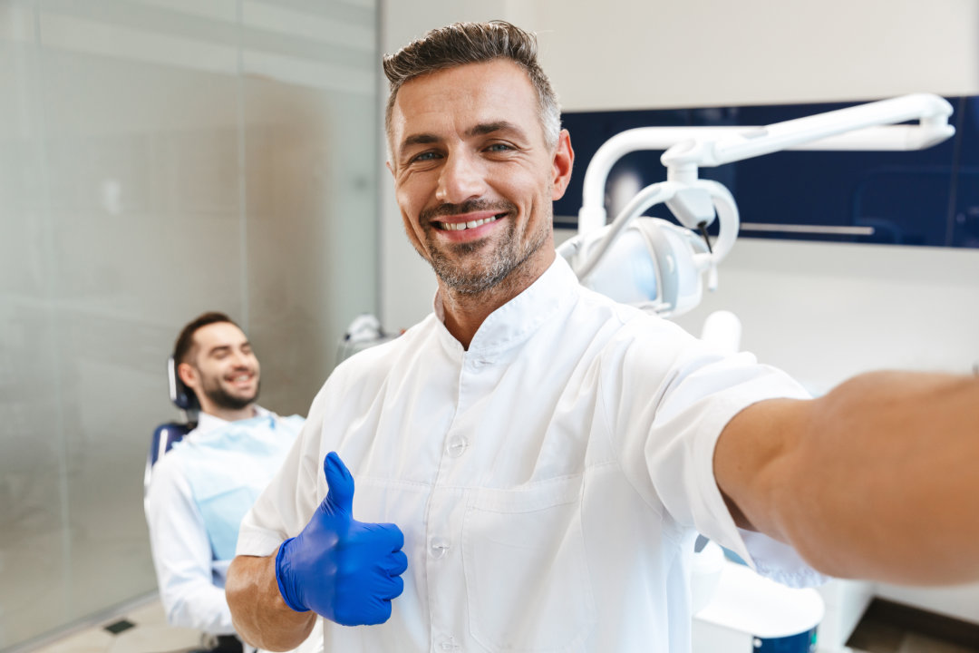 Image of a handsome happy young man doctor in medical dentist center showing thumbs up take a selfie by camera.